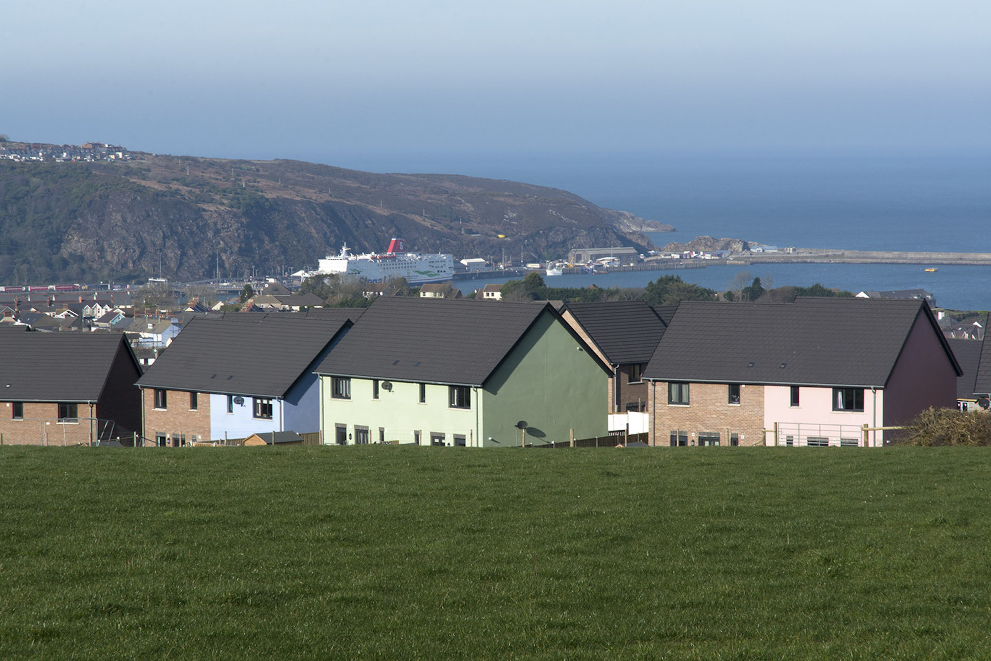 Grass to foreground leading to housing development with colourful render and sea blurred in horizon line